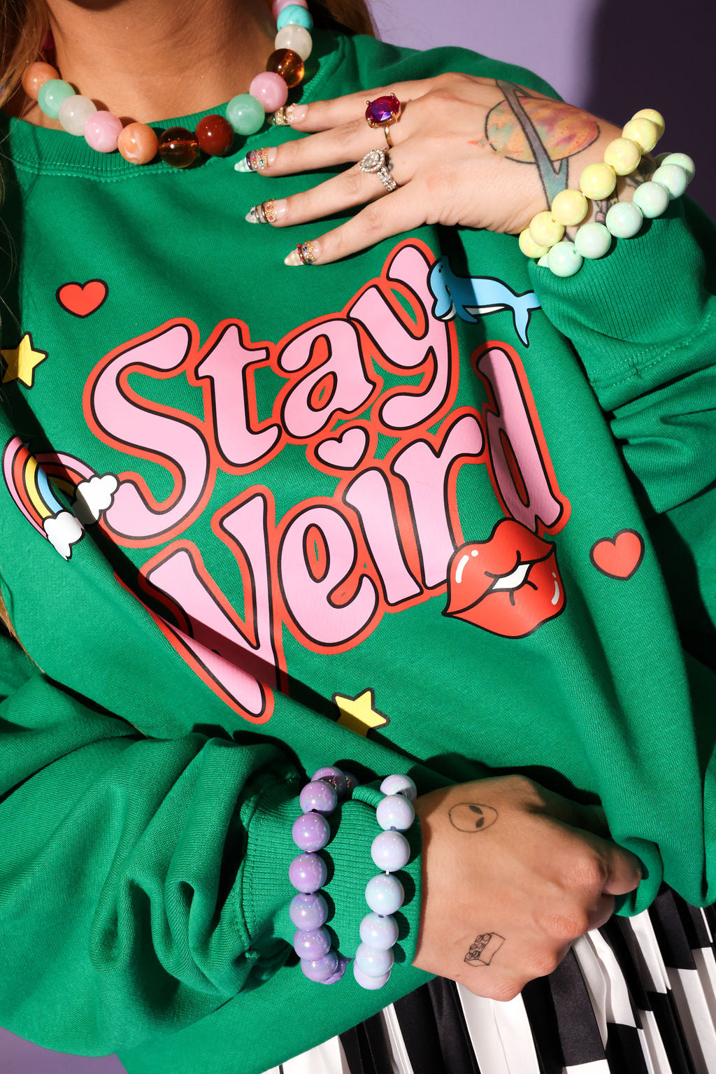 Person wearing a green sweatshirt with 'Stay Weird!' text, surrounded by colorful jewelry