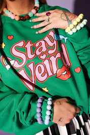 Person wearing a green sweatshirt with 'Stay Weird!' text, surrounded by colorful jewelry