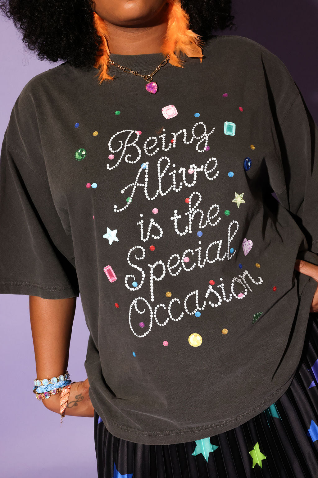 Woman standing on a ladder wearing a gray graphic t-shirt with text 'Being Alive is the Special Occasion' and star-patterned skirt against a purple background, close up frontal view