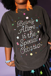 Woman standing on a ladder wearing a gray graphic t-shirt with text 'Being Alive is the Special Occasion' and star-patterned skirt against a purple background, close up frontal view