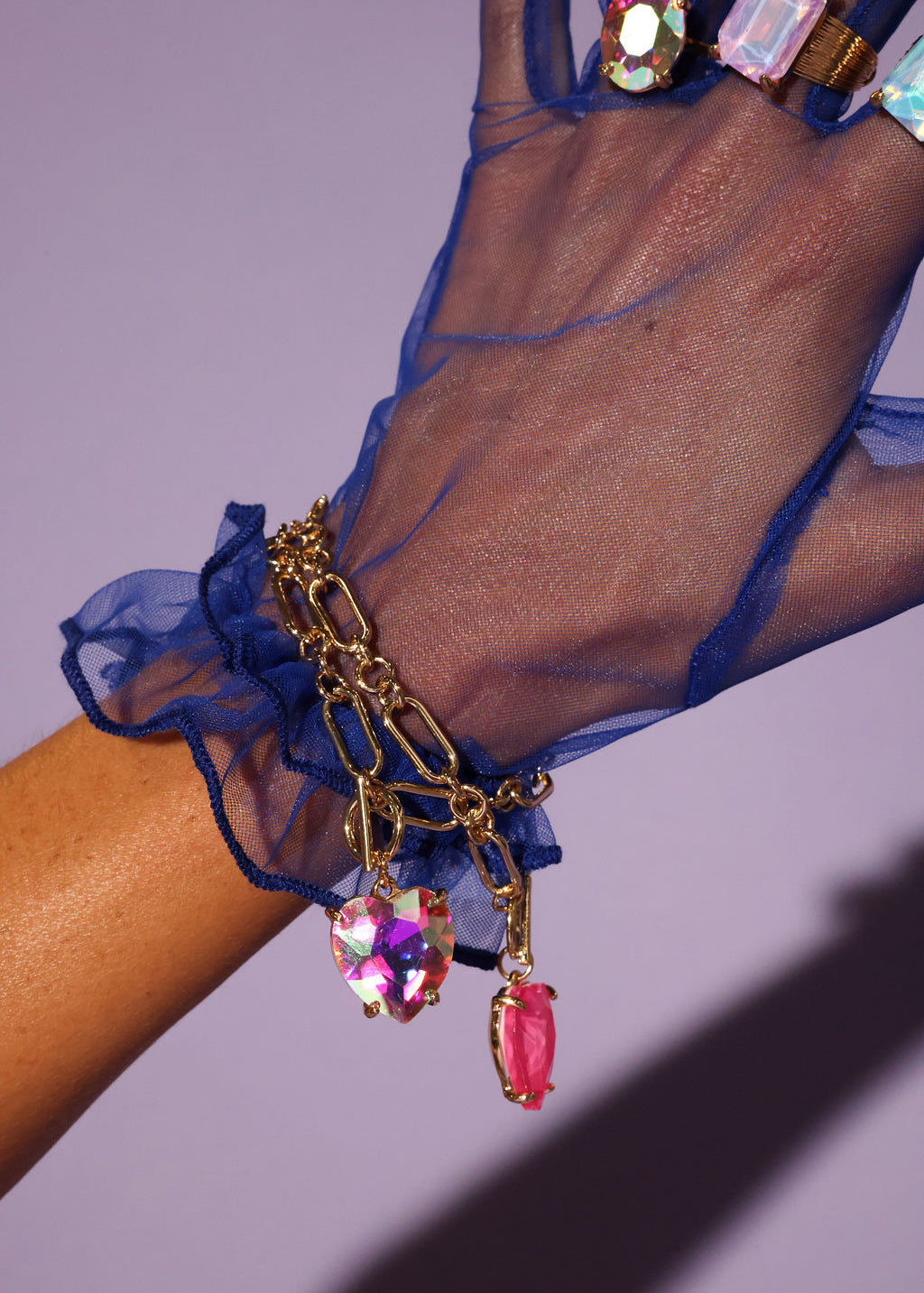 Hand wearing a blue sheer glove with a gold bracelet featuring colorful gemstones on a purple background