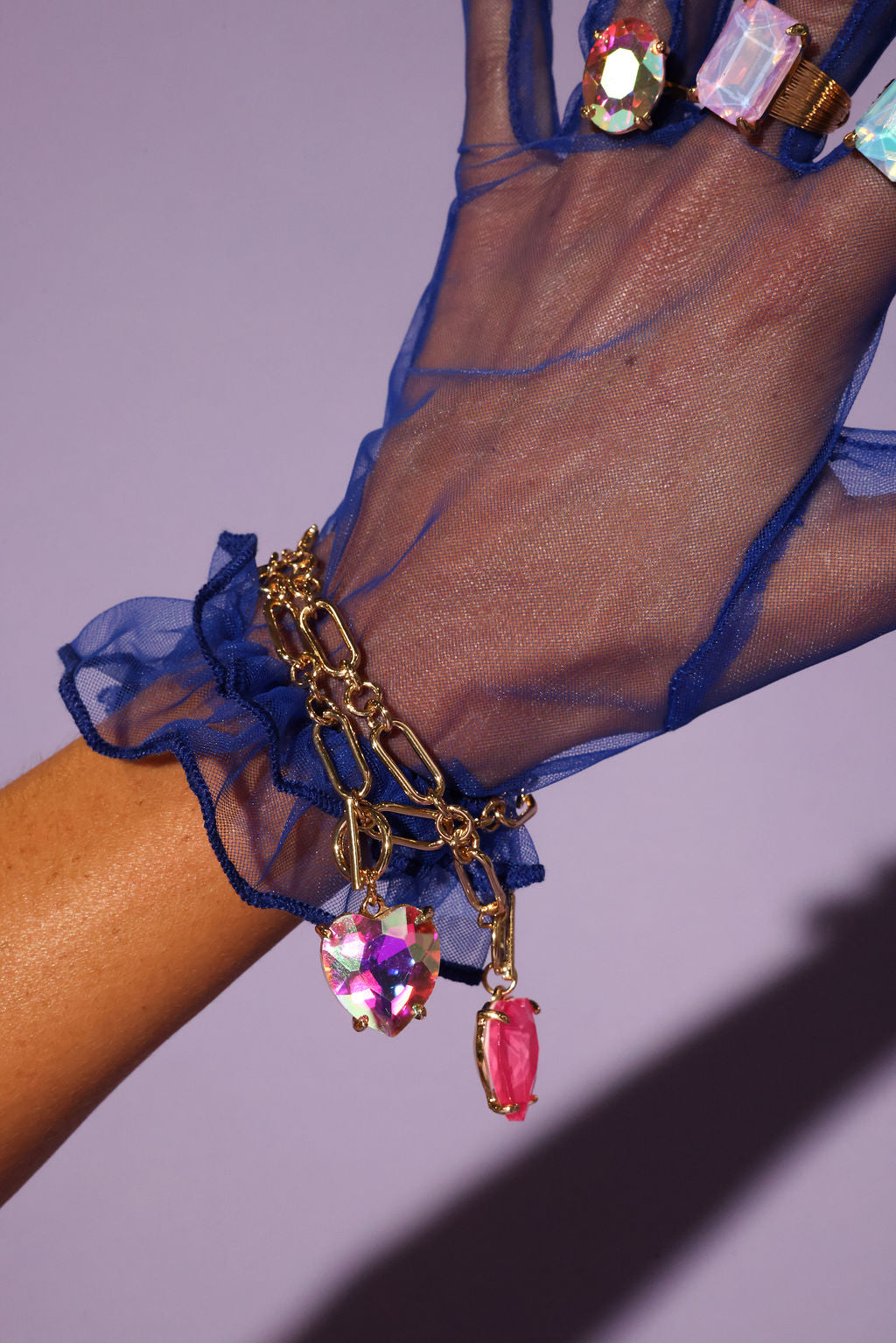 Hand wearing a blue sheer glove with a gold bracelet featuring colorful gemstones on a purple background
