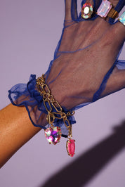 Hand wearing a blue sheer glove with a gold bracelet featuring colorful gemstones on a purple background