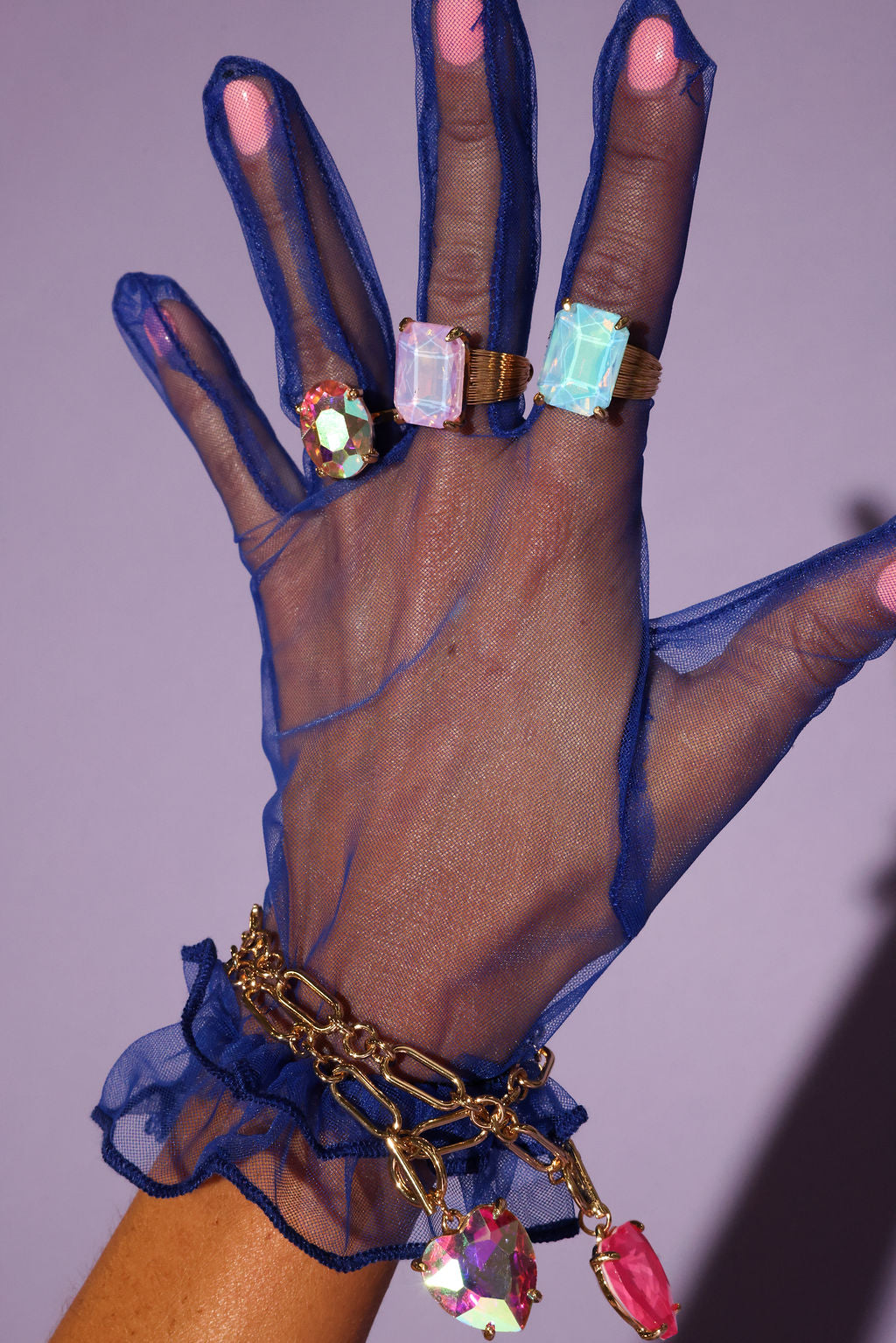 Hand wearing a blue sheer glove with colorful gemstone rings and bracelets against a purple background