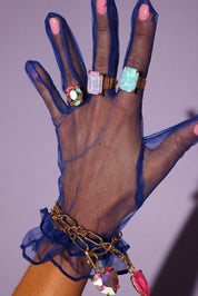 Hand wearing a blue sheer glove with colorful gemstone rings and bracelets against a purple background