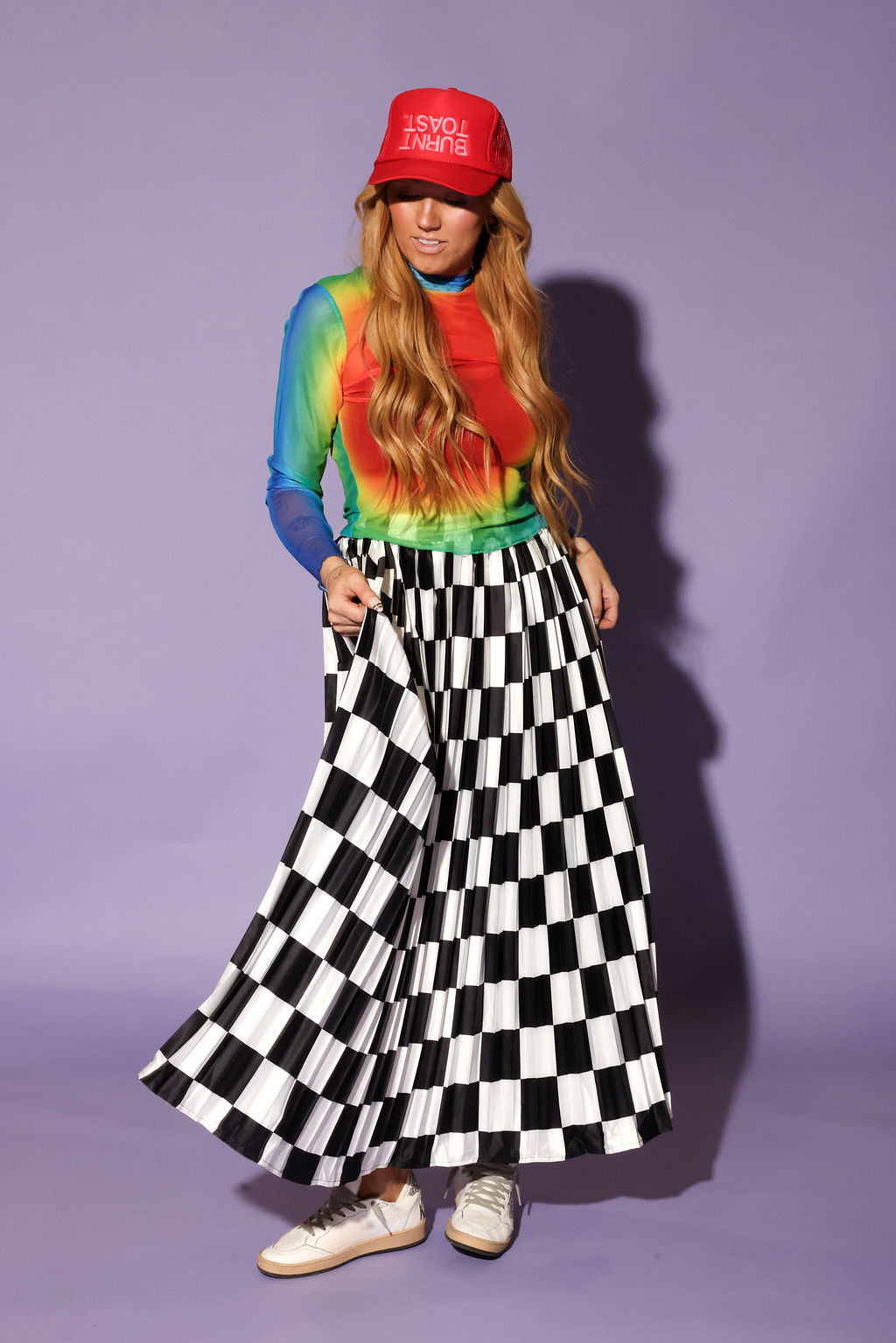 A woman modeling a vibrant rainbow tie-dye long-sleeve top, a red baseball cap with white text, and a black and white checkered pleated maxi skirt, standing against a solid lavender studio background, frontal view
