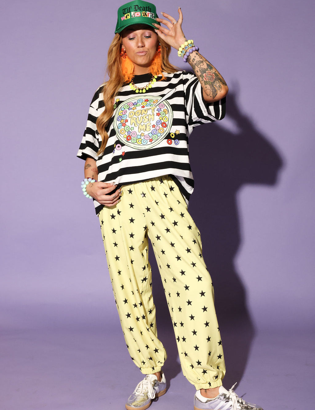 Model wearing a striped shirt, yellow pants with star patterns, and a green cap against a purple background, frontal view