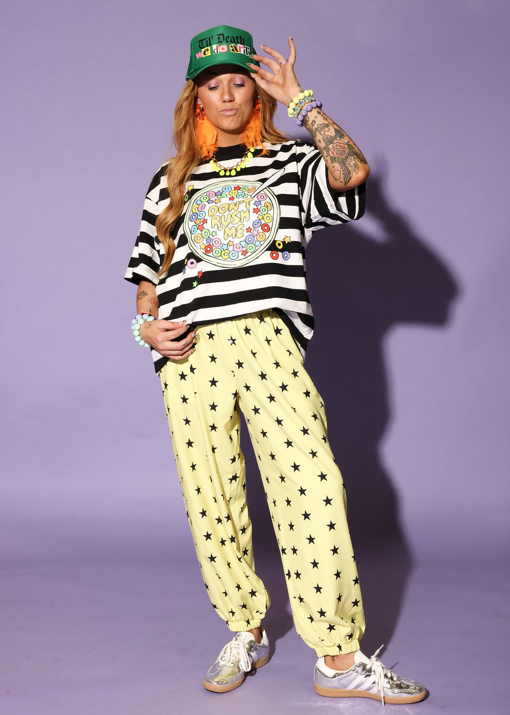 Model wearing a striped shirt, yellow pants with star patterns, and a green cap against a purple background, frontal view