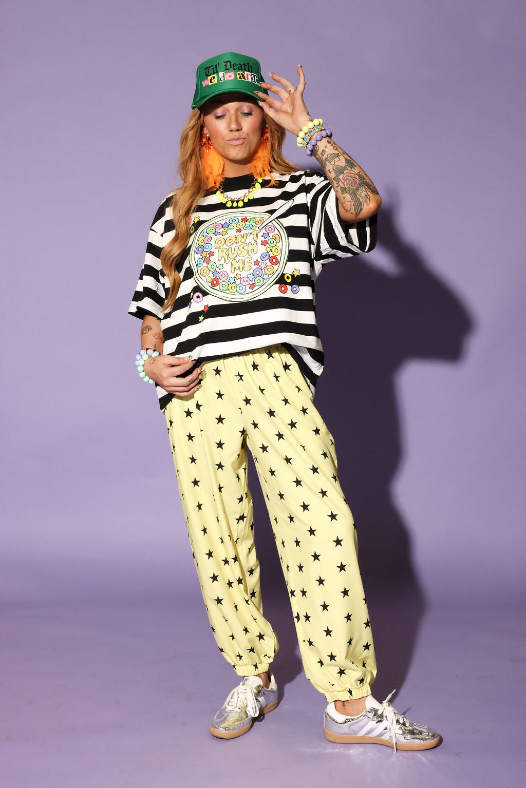 Model wearing a striped shirt, yellow pants with star patterns, and a green cap against a purple background, frontal view