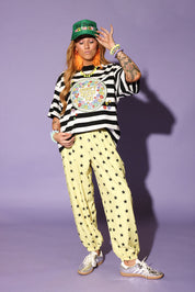 Model wearing a striped shirt, yellow pants with star patterns, and a green cap against a purple background, frontal view