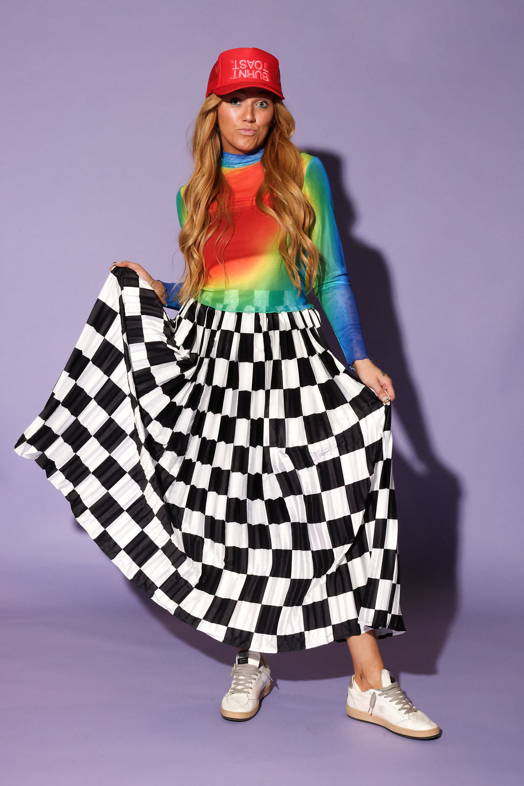 A woman modeling a vibrant rainbow tie-dye long-sleeve top, a red baseball cap with white text, and holding a black and white checkered pleated maxi skirt, standing against a solid lavender studio background, frontal view