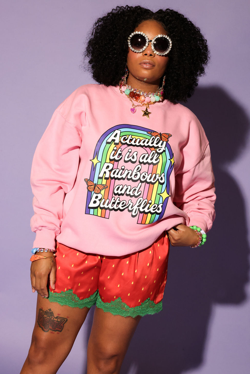 Person wearing a pink sweatshirt with a rainbow design and text, red shorts with green trim, and sunglasses on a purple background