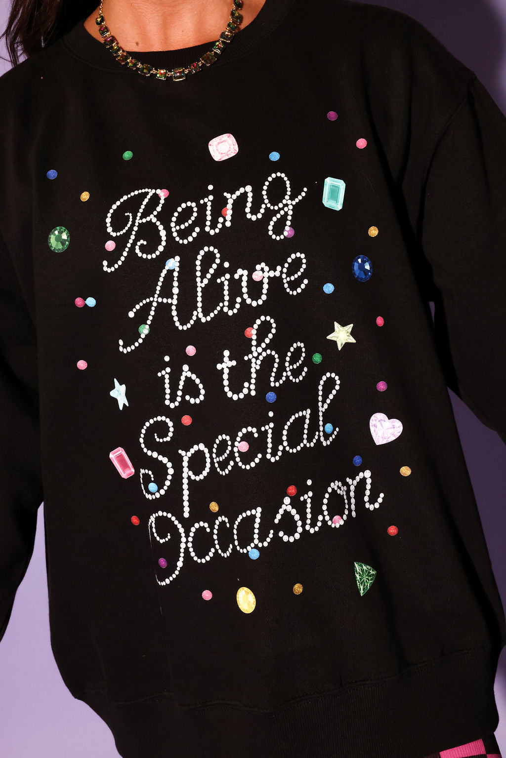 Black sweatshirt with colorful text and rhinestone embellishments on a purple background, close up frontal view