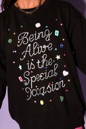 Black sweatshirt with colorful text and rhinestone embellishments on a purple background, close up frontal view