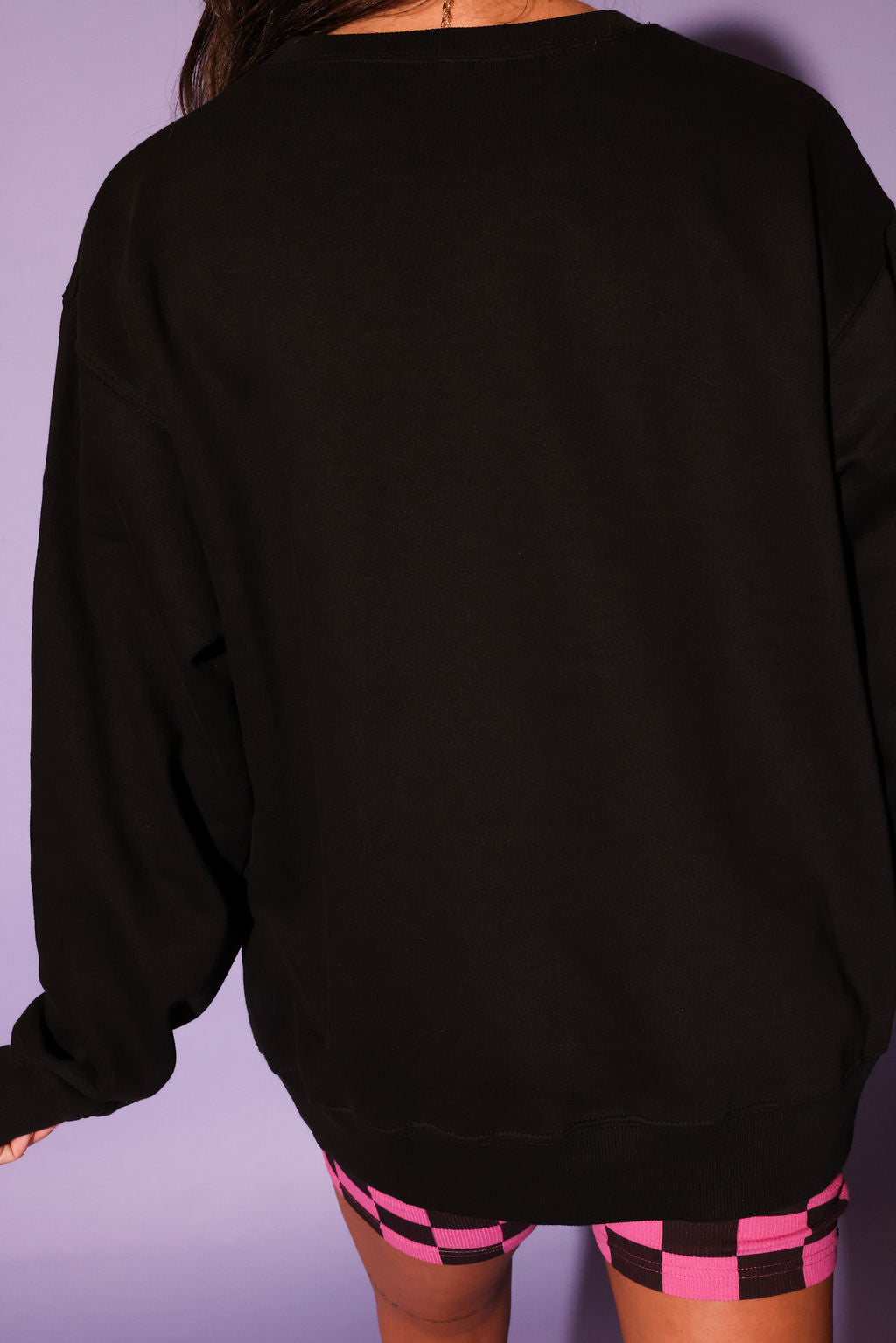 Person wearing a black oversized sweatshirt and checkered shorts against a purple background, back view
