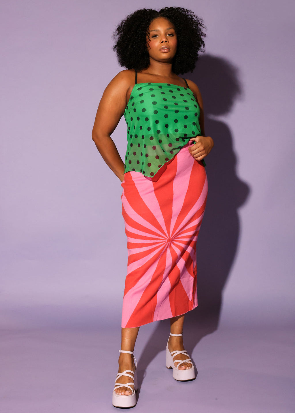 Woman wearing a green top with black polka dots and a red and pink striped skirt on a purple background, frontal view