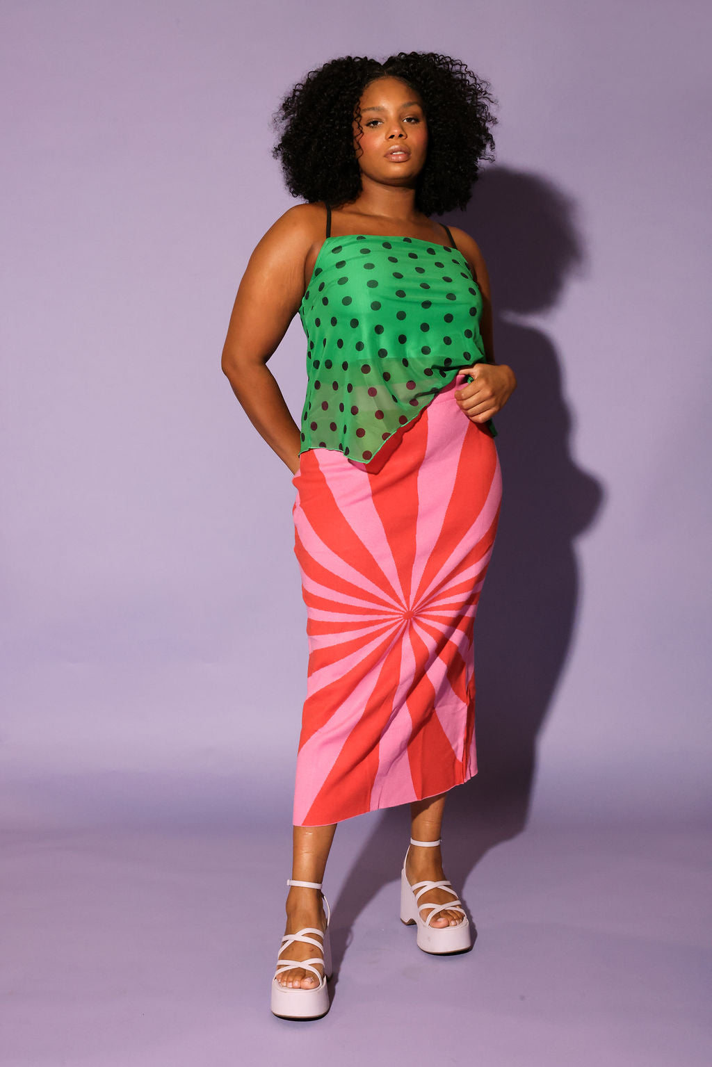 Woman wearing a green top with black polka dots and a red and pink striped skirt on a purple background, frontal view