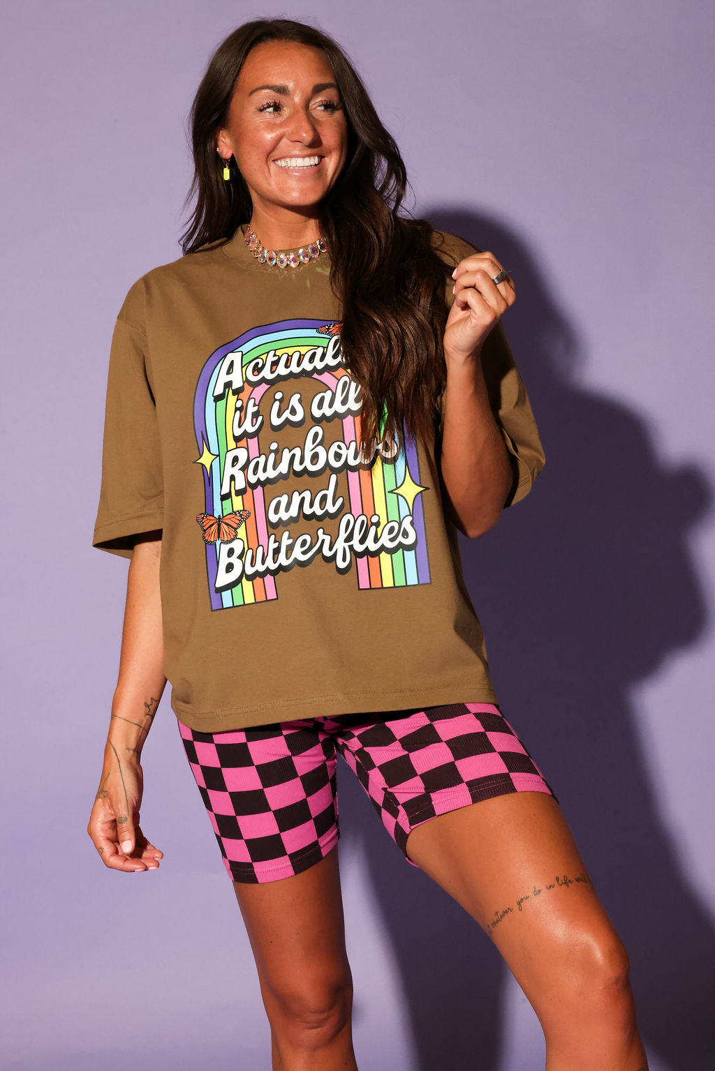 Woman wearing a brown t-shirt with colorful text and pink checkered shorts against a purple background, frontal view
