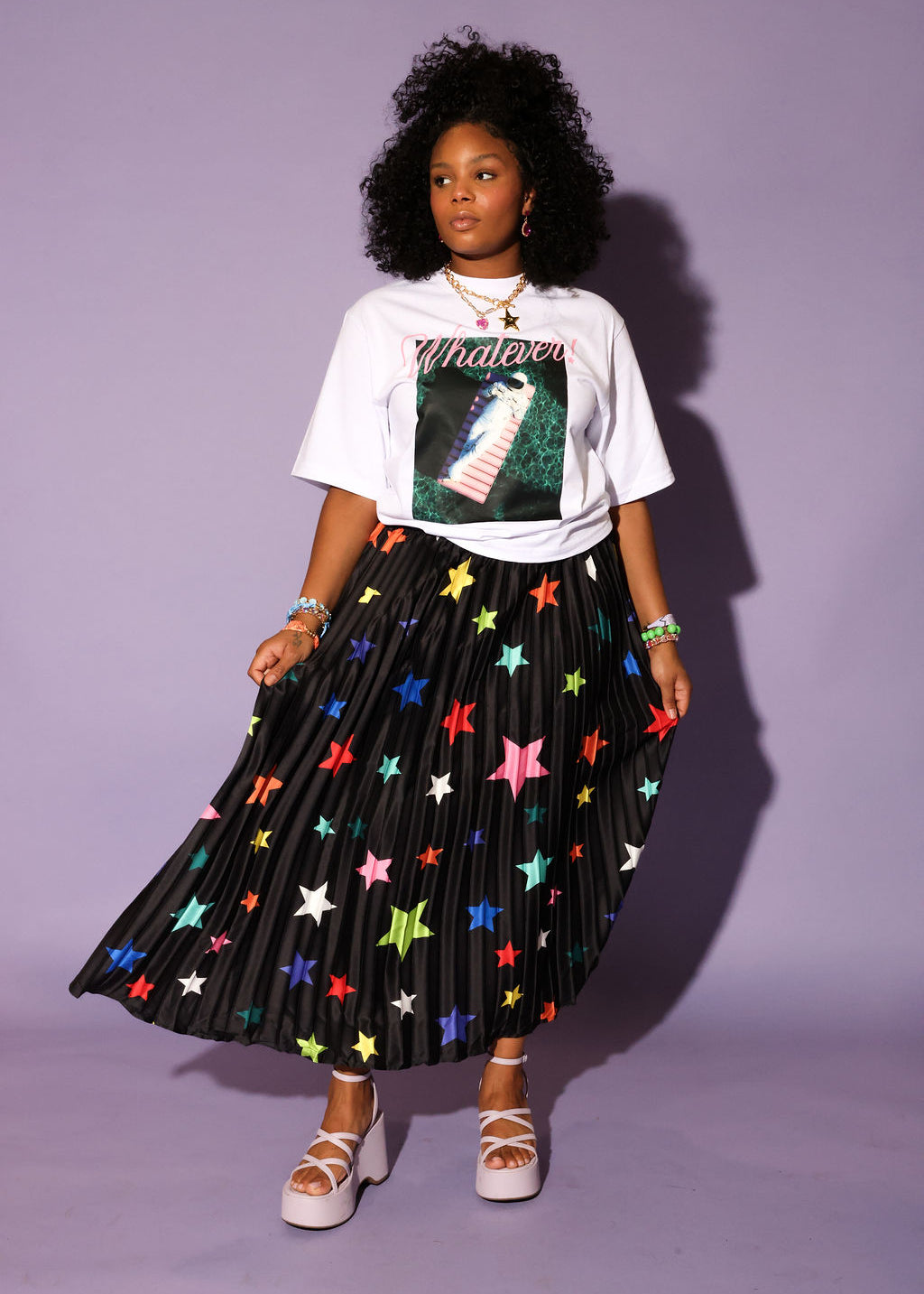 Model wearing a white graphic tee shirt, a colorful star patterned black plisse long skirt, pink wedges, on an purple background, frontal view