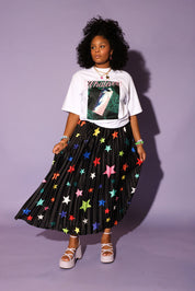 Model wearing a white graphic tee shirt, a colorful star patterned black plisse long skirt, pink wedges, on an purple background, frontal view