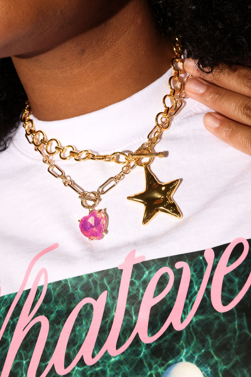Gold chain necklace with star and pink charm worn by a person, with a white shirt and colorful text in the background