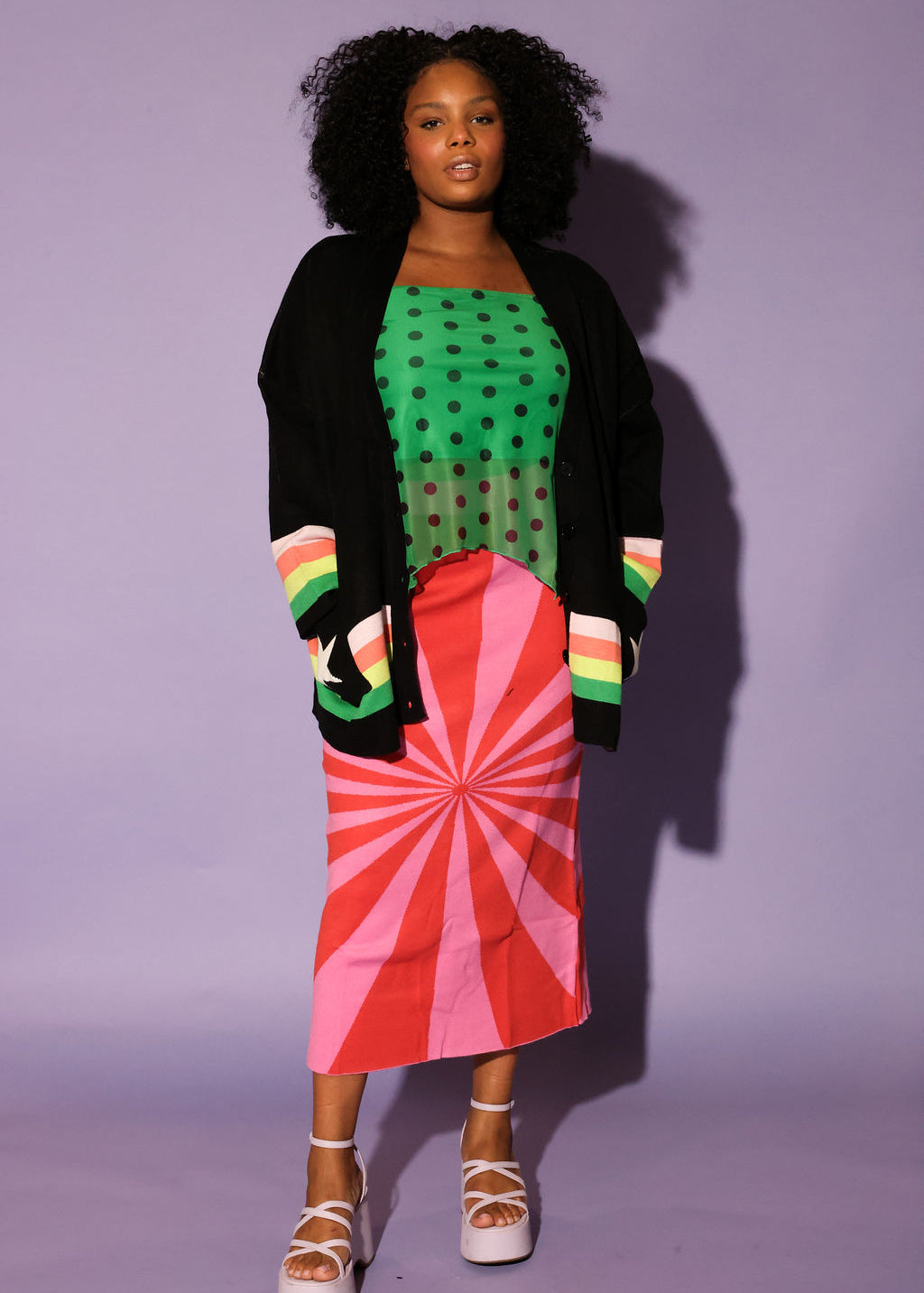 Woman wearing a green polka dot top, black cardigan, red and pink skirt, and colorful striped shoes against a purple background, frontal view