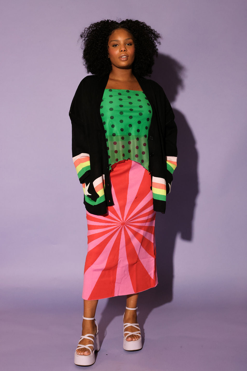 Woman wearing a green polka dot top, black cardigan, red and pink skirt, and colorful striped shoes against a purple background, frontal view