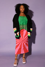 Woman wearing a green polka dot top, black cardigan, red and pink skirt, and colorful striped shoes against a purple background, frontal view