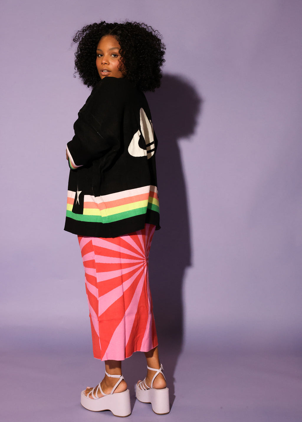 Person wearing a black sweater with colorful stripes and a pink skirt against a purple background, side view