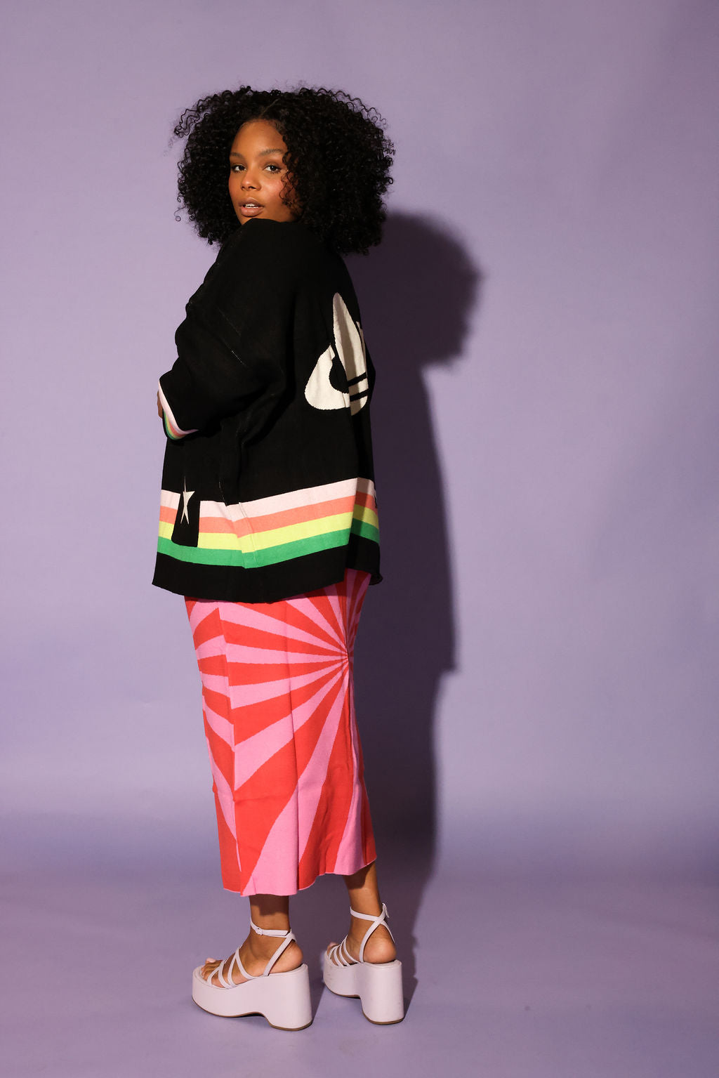 Person wearing a black sweater with colorful stripes and a pink skirt against a purple background, side view