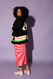 Person wearing a black sweater with colorful stripes and a pink skirt against a purple background, side view