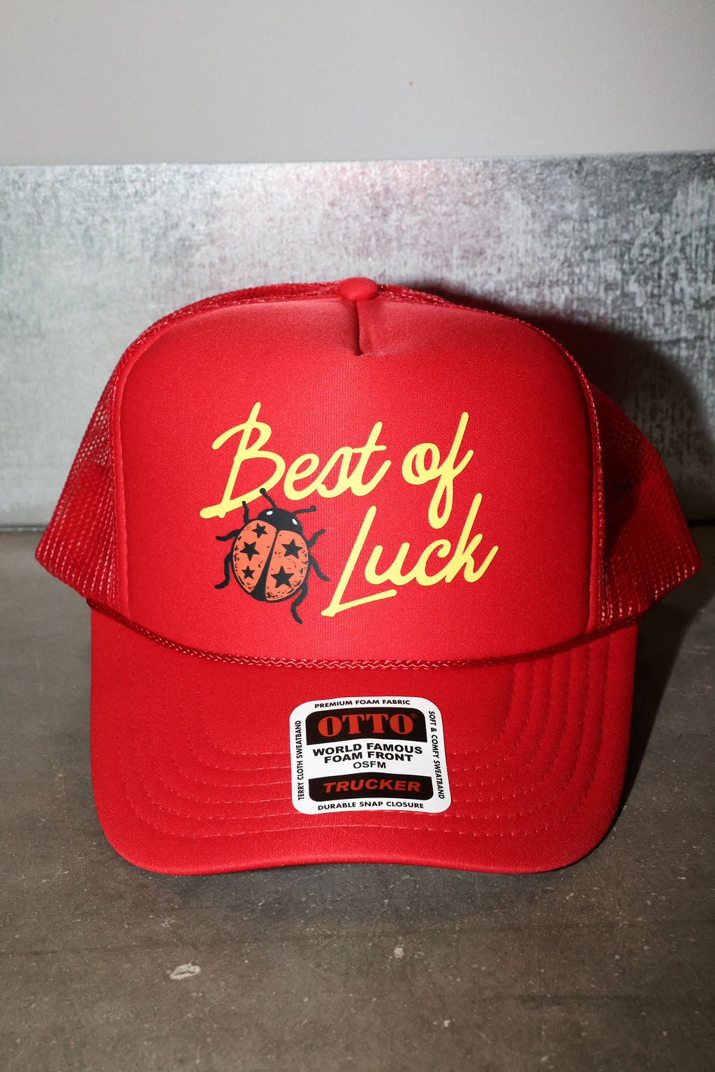A red trucker hat with 'Best of Luck' and a ladybug graphic embroidered on the front, featuring a visible OTTO brand patch on the brim