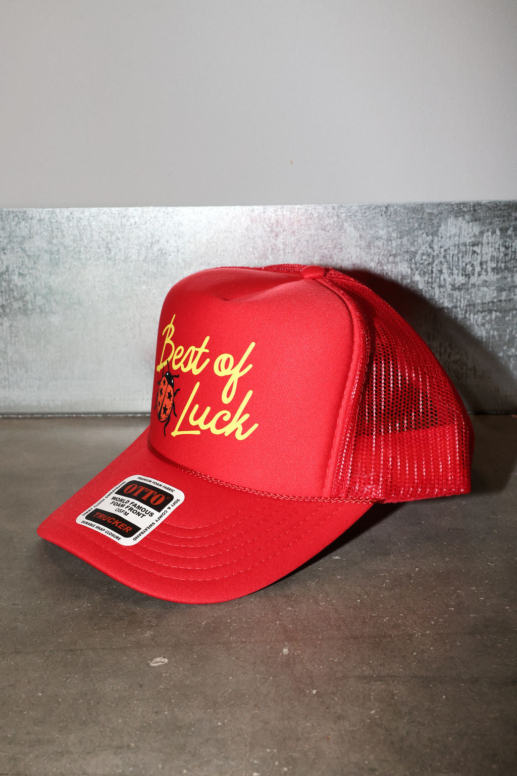 A red trucker hat with yellow embroidered text reading 'Best of Luck' and a small devil graphic, shown on a gray concrete surface against a gray wall