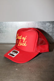 A red trucker hat with yellow embroidered text reading 'Best of Luck' and a small devil graphic, shown on a gray concrete surface against a gray wall