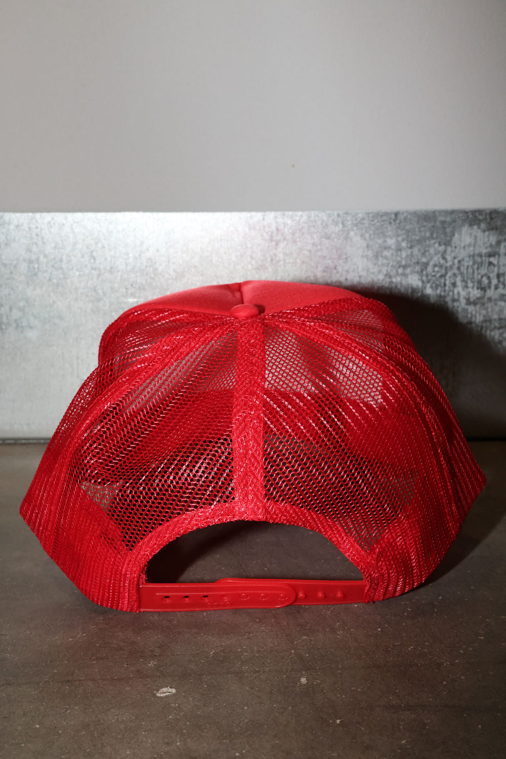 A bright red trucker-style cap with a red mesh back and red adjustable strap, photographed against a gray wall and dark floor