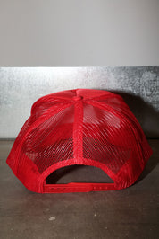 A bright red trucker-style cap with a red mesh back and red adjustable strap, photographed against a gray wall and dark floor