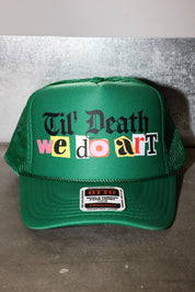 A green trucker hat with a colorful graphic reading 'Til' Death we do art' on the front, photographed against a gray textured wall