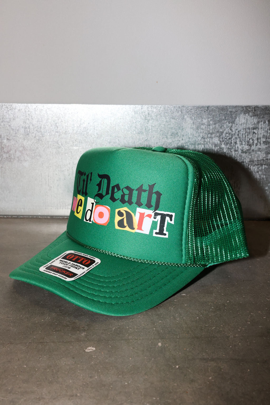 A green trucker hat with a mesh back, featuring the text 'Til Death do art' on the front panel. The letters 'e', 'o', and 'a' are styled in red, yellow, and orange colors, sticker are in black and white