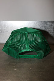 A green trucker-style cap with a mesh back, photographed from the back against a textured gray wall and concrete floor