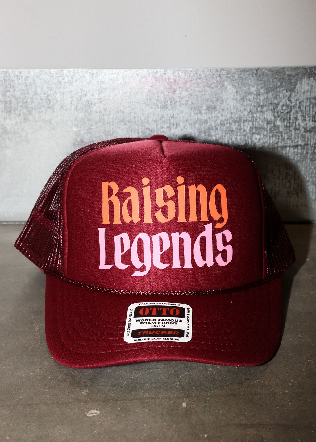 A maroon trucker hat with 'Raising Legends' printed on the front in orange and pink, featuring a white brand patch on the brim