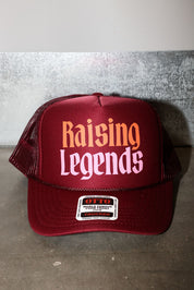 A maroon trucker hat with 'Raising Legends' printed on the front in orange and pink, featuring a white brand patch on the brim
