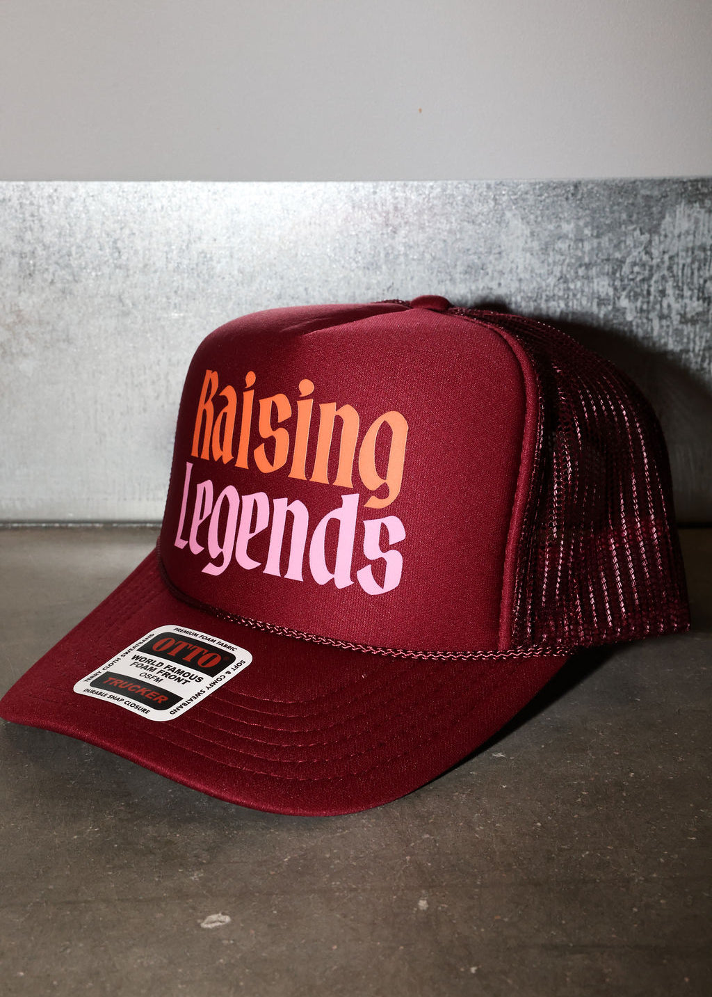 A maroon trucker hat with 'Raising Legends' printed on the front in orange and white, featuring a maroon mesh back and a visible sticker on the brim