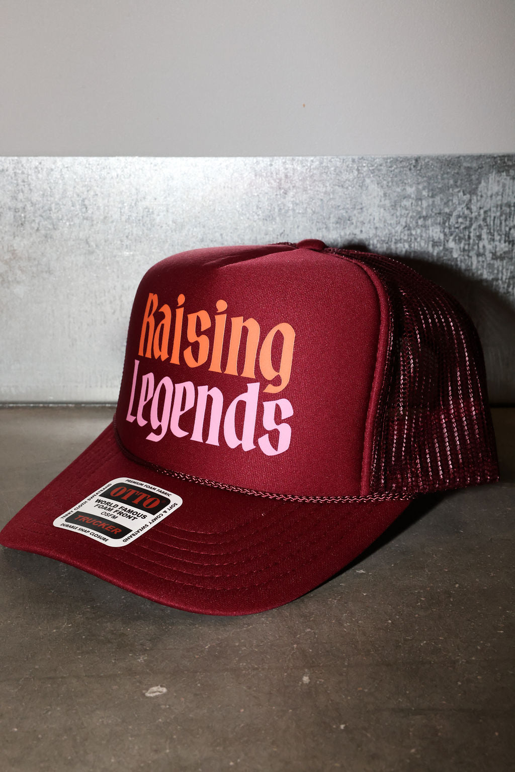 A maroon trucker hat with 'Raising Legends' printed on the front in orange and white, featuring a maroon mesh back and a visible sticker on the brim