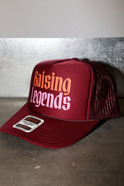 A maroon trucker hat with 'Raising Legends' printed on the front in orange and white, featuring a maroon mesh back and a visible sticker on the brim