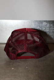 A burgundy trucker-style baseball cap with a mesh back and adjustable strap, photographed against a textured gray wall and concrete floor