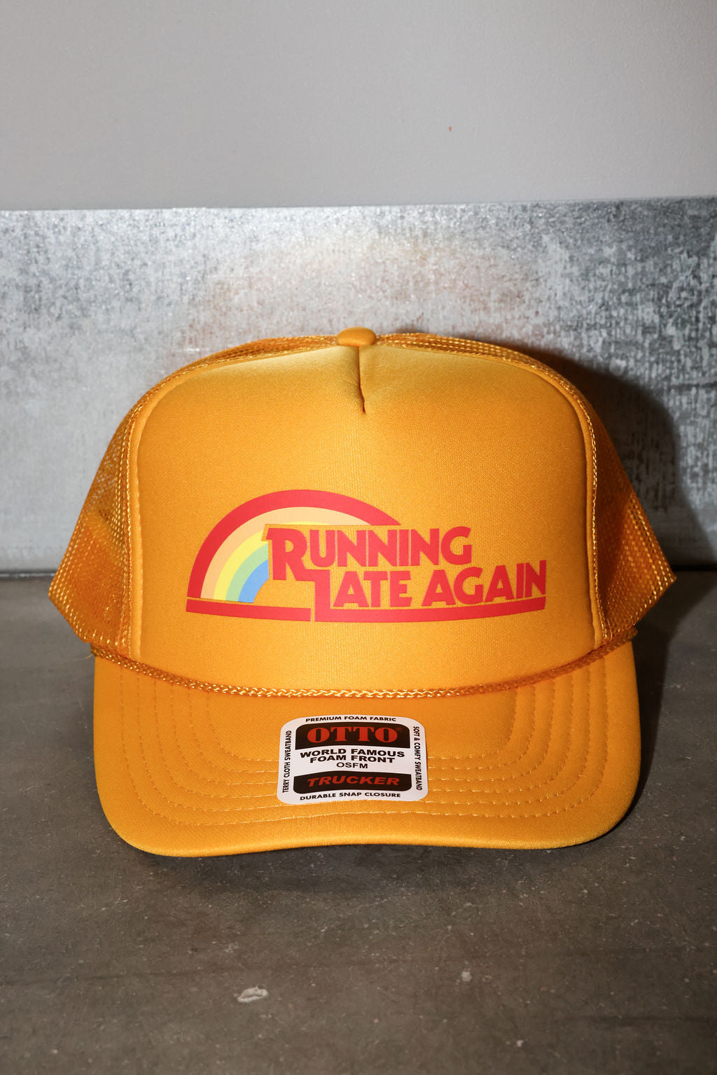 Mustard trucker hat with the text 'RUNNING LATE AGAIN' in red with a rainbow, frontal view