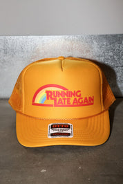 Mustard trucker hat with the text 'RUNNING LATE AGAIN' in red with a rainbow, frontal view