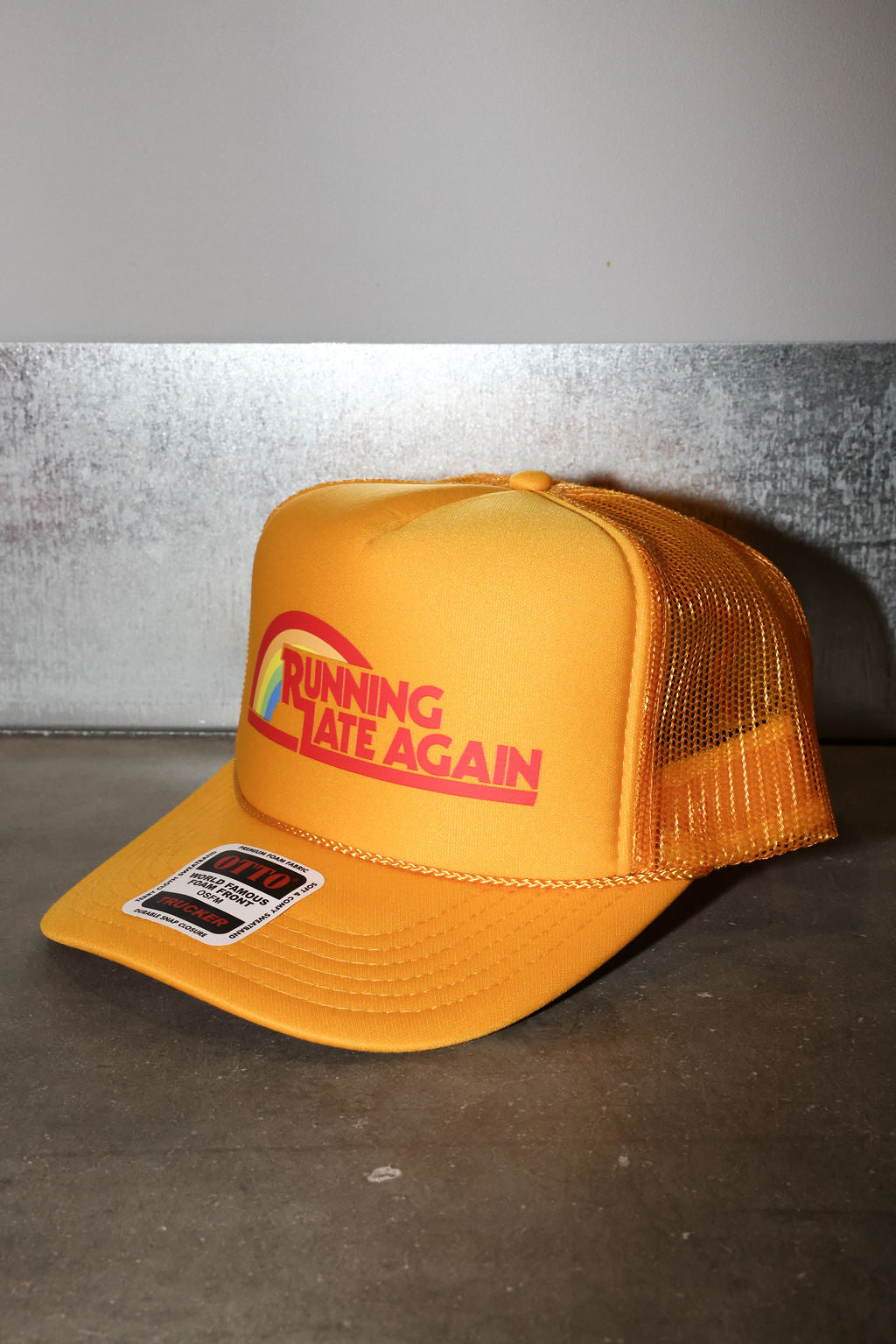 A yellow-orange mesh-back trucker hat with a curved brim, featuring a red and white graphic on the front that reads 'RUNNING LATE AGAIN' and a small sticker on the brim