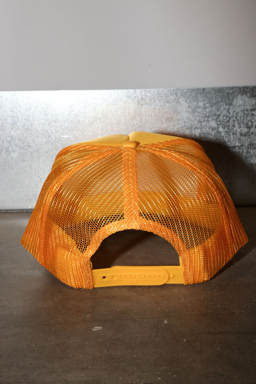 A mustard mesh trucker hat with a solid orange fabric front panel and adjustable strap, photographed against a gray and dark background.