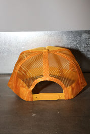 A mustard mesh trucker hat with a solid orange fabric front panel and adjustable strap, photographed against a gray and dark background.